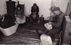 A black-and-white photo of two women and a man weaving baskets