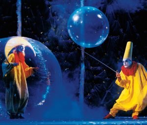 One clown inside a plastic bubble; another clown bounces a large bubble on a stick.