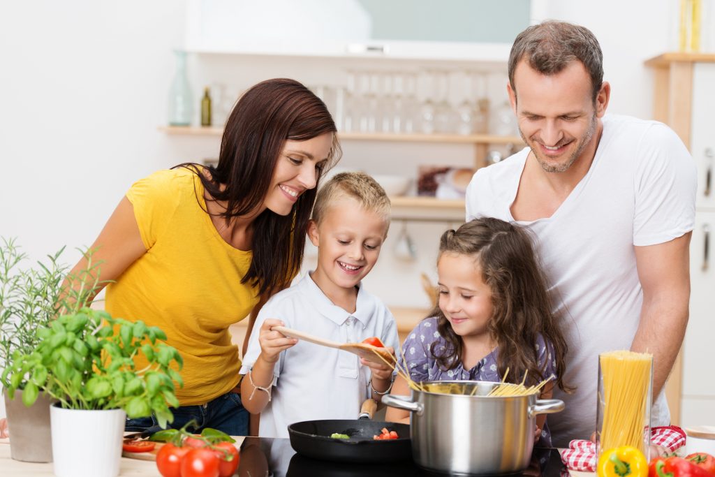 A mother, father, and two young children standing by the stove, cooking pasta and pasta sauce