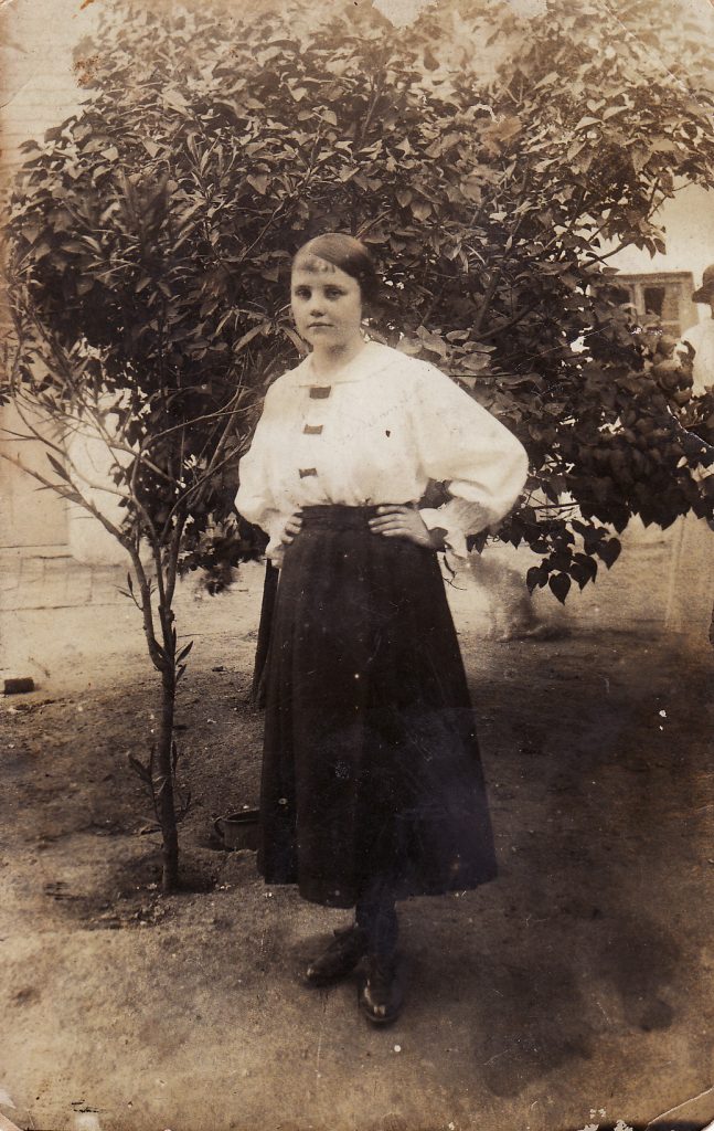 A young teenage girl wearing a white blouse and dark skirt and standing in front of a small tree. The photo is from the 1910s