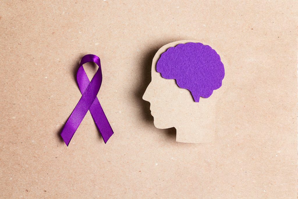 A purple ribbon and a cutout of the profile of a head, with a purple brain. Both represent epilepsy, including absence seizures.