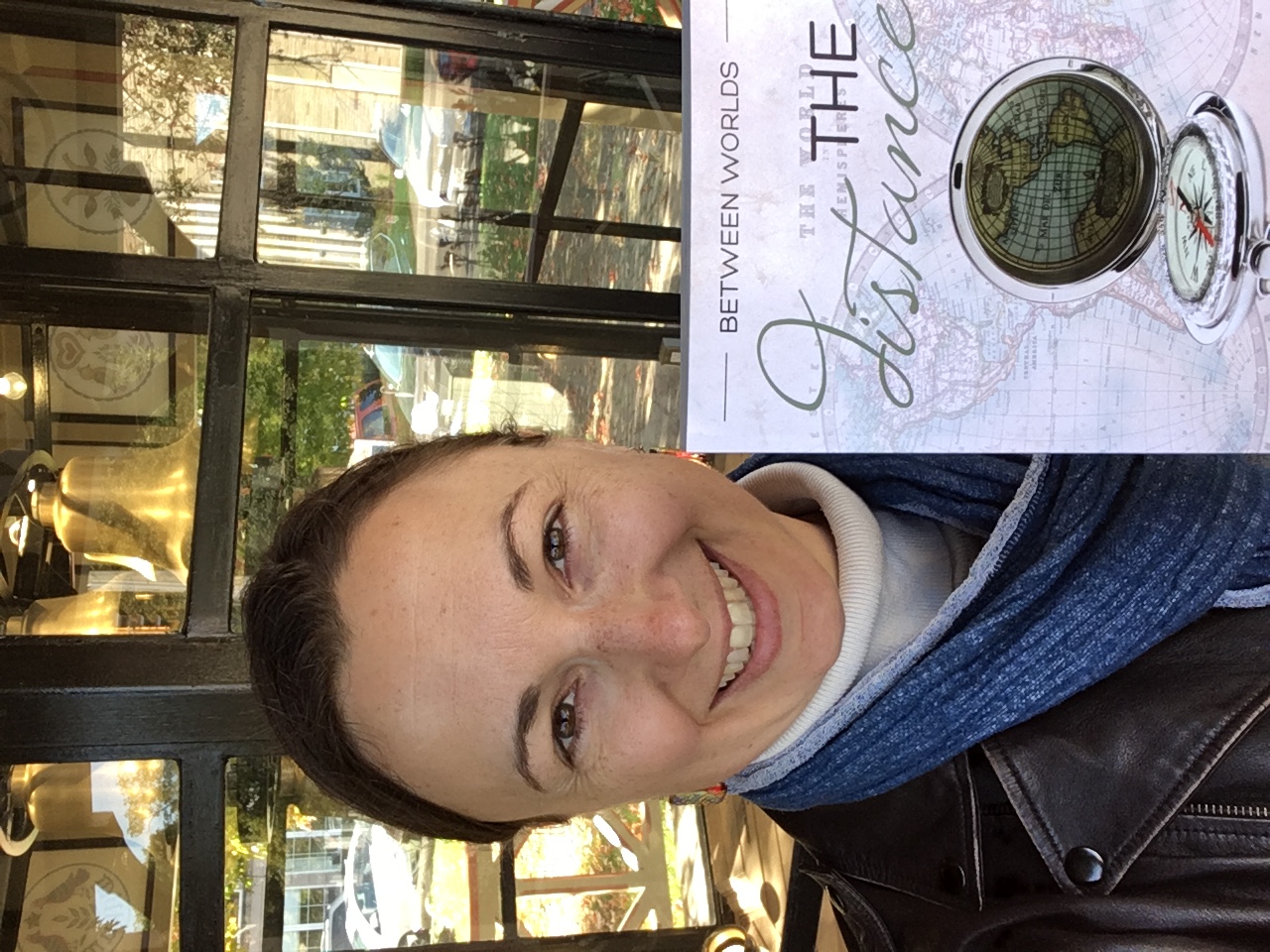 Lori Wolf-Heffner holding a copy of Tea Shop for Two in front of the rusty bell sculpture mentioned in the book