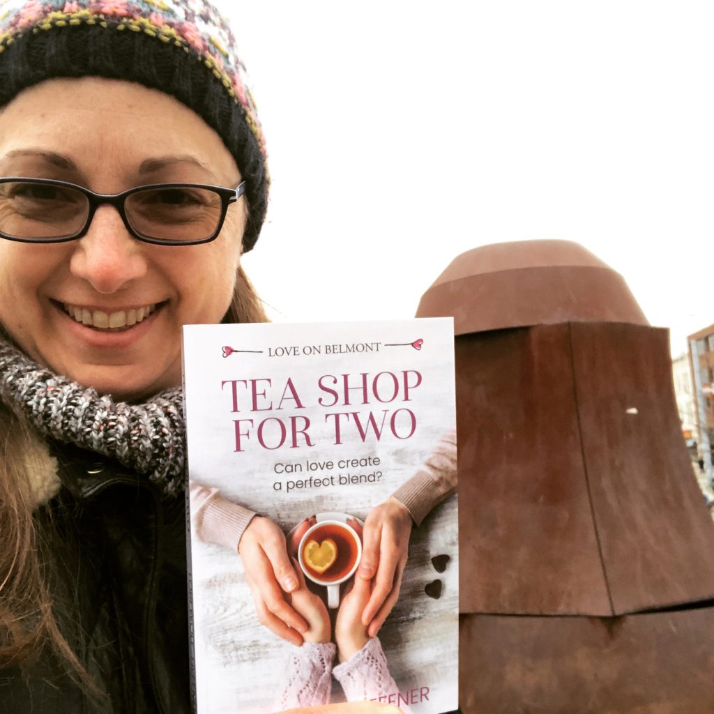 Lori Wolf-Heffner in a toque and winter jacket holding a copy of Tea Shop for Two in front of a rusty bell sculpture in Waterloo, Ontario.