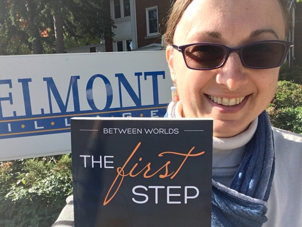Lori Wolf-Heffner holds a copy of Between Worlds 3: The First Step in front of the Belmont Village sign.