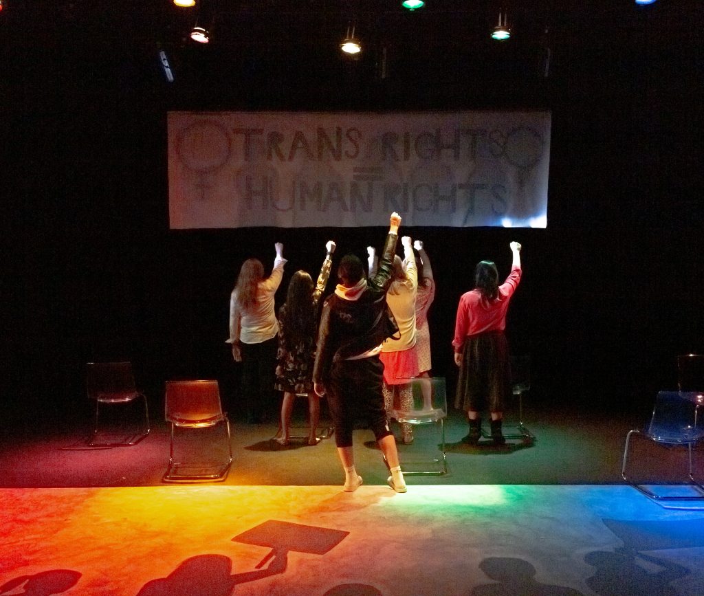 Actors on stage facing the back, raising their hands in fists in support of a sign that says, "Trans rights = human rights."
