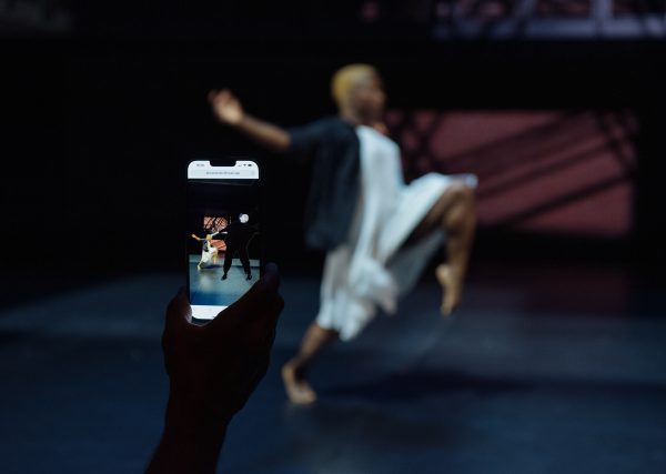 Background: blurred-out woman taking a large step on the stage. Foreground: smartphone of the woman on the stage, with the image of another woman superimposed beside her, but larger.