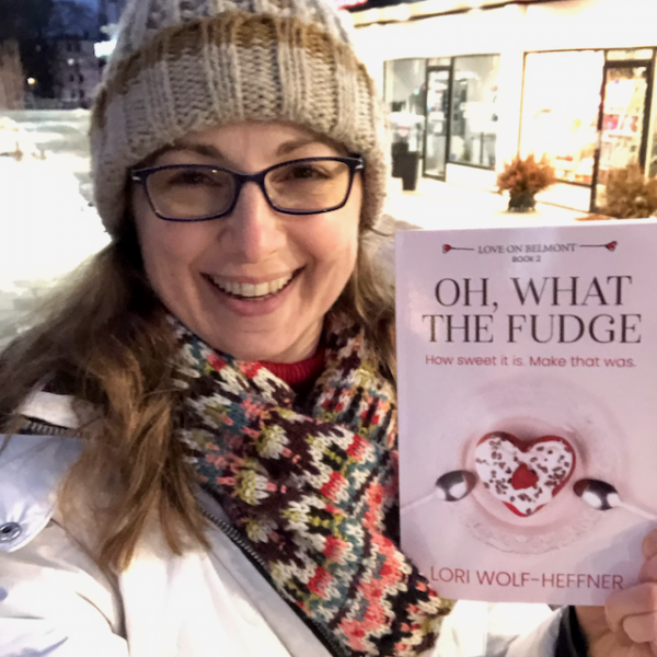 Lori is dressed in outdoor winter clothes and holding a copy of "Oh, What the Fudge." Shops from Belmont Village Kitchener are behind her.