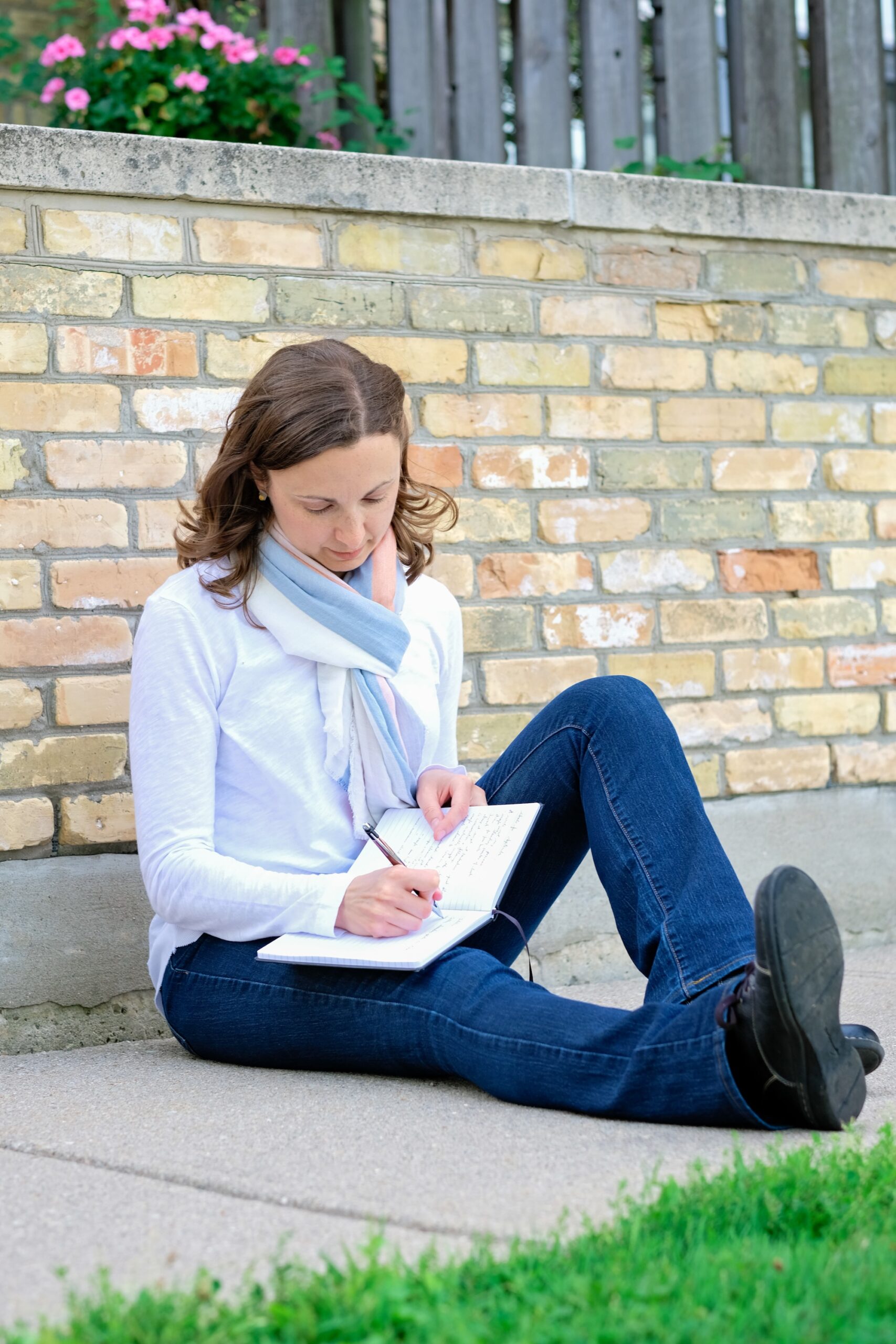 Lori Wolf-Heffner is sitting against a yellow brick wall, writing in a journal.